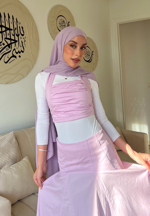 Nainen, jolla on vaalean violetti huntu, sopiva yläosa ja hame, seisoo sisätiloissa sohvalla, jonka takana seinällä on arabialaista kalligrafiaa.