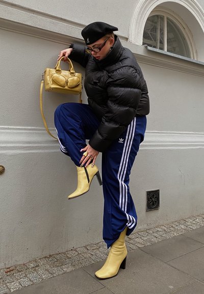 Chaqueta acolchada negra, pantalones de chándal azules de Adidas con rayas blancas, botas de tacón amarillas y un pequeño bolso amarillo. Texturas suaves, colores llamativos.