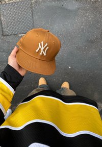 Cappellino da baseball marrone con logo "NY" ricamato in bianco. Maglietta a righe nera e gialla visibile in primo piano. Pavimento scuro sottostante.