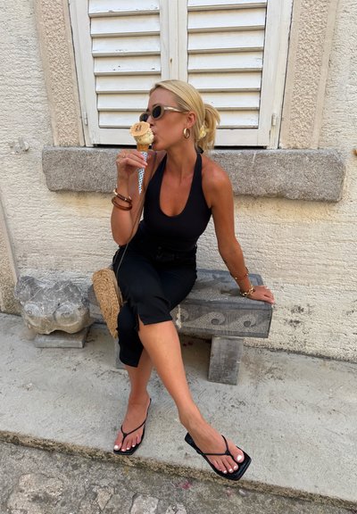 Top negro de halter y pantalones negros a medida, sosteniendo un cono de helado. Sandalias negras planas; una bolsa tejida descansa a su lado en un banco de piedra.