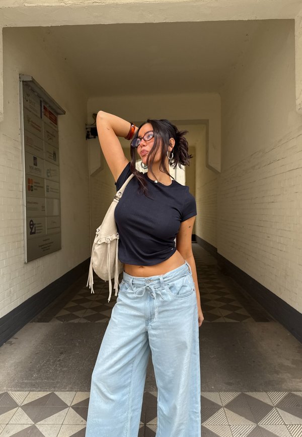 T-shirt nero aderente, jeans in denim a gamba larga di colore azzurro chiaro, borsa a spalla bianca con dettagli in frange e orecchini decorativi. Ambientazione interna.