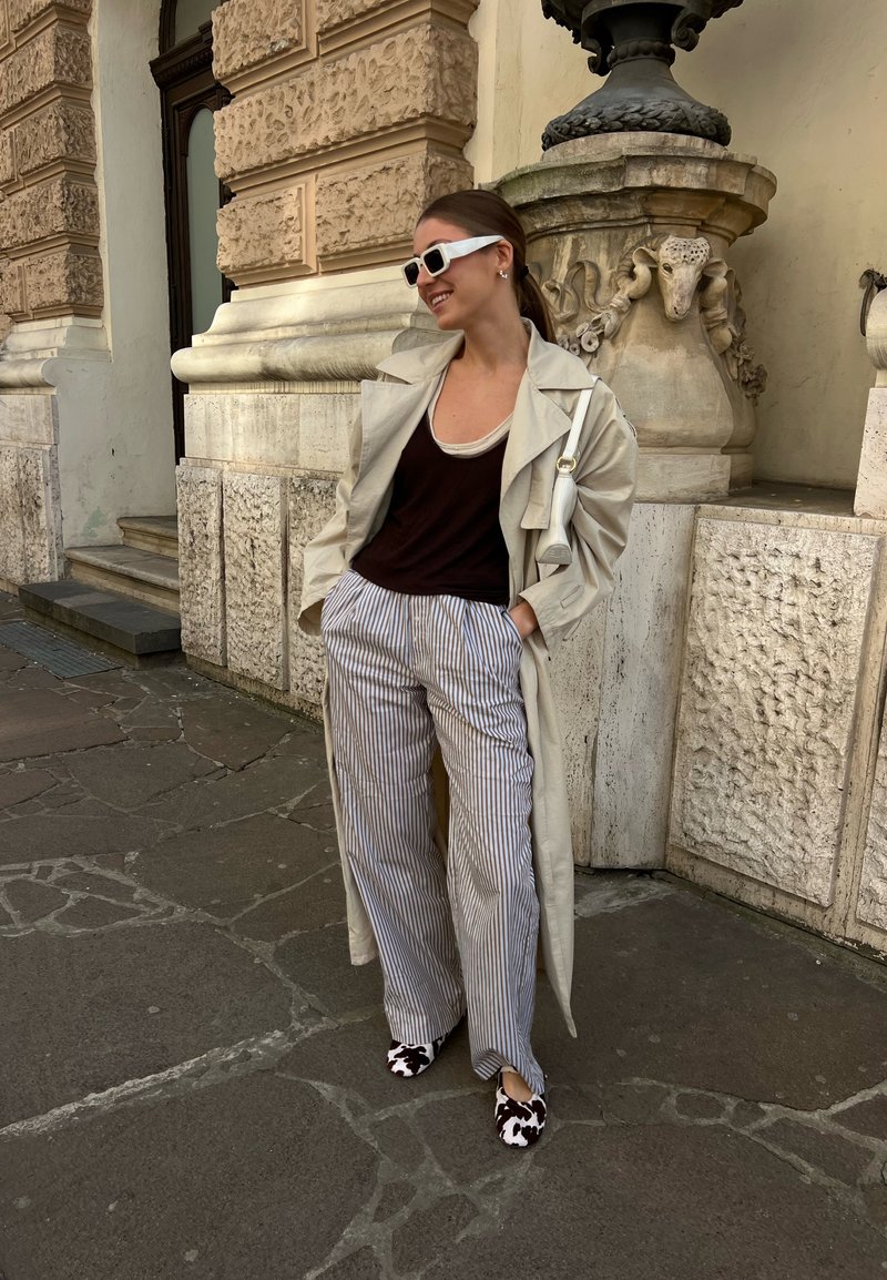 Femme portant un trench-coat beige, un haut noir, un pantalon large à rayures, des lunettes de soleil blanches et des chaussures à motifs, appuyée contre un mur de pierre orné à l'extérieur.