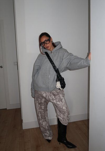 Mujer con sudadera gris oversize, pantalones estampados, botas negras de tacón, gafas grandes y un bolso bandolera negro con un llavero blanco, apoyada contra una pared.