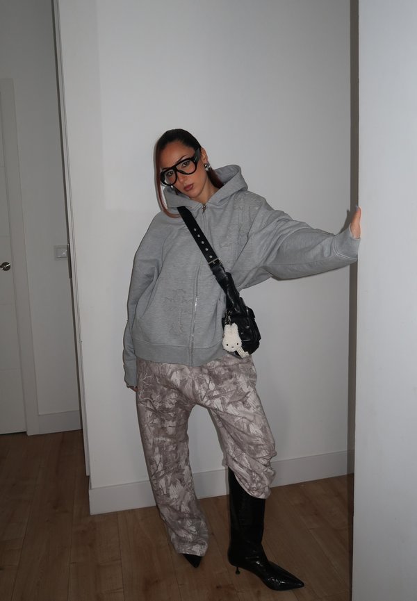 Naine, kes kannab liibuvat halli kapuutsi, mustrilisi pükse, musti kontsakingi, suuri prille ja musta ristiõlakotti valge võtmehoidjaga, toetub seinale.