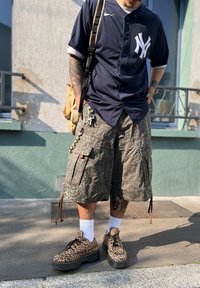Granatowa koszulka baseballowa z białym logo, wzorzyste zielone szorty cargo i brązowe buty na platformie w wzór lamparta, w zestawie z białymi skarpetkami.