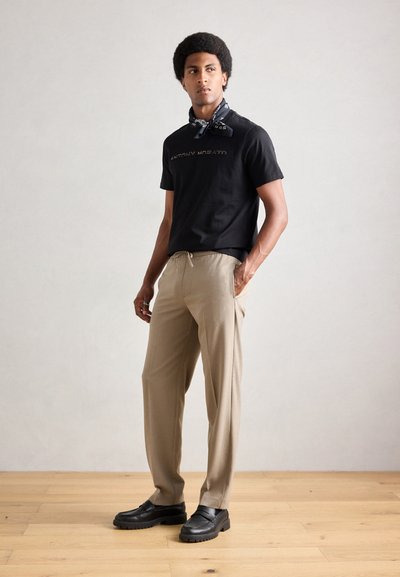 Camiseta de algodón negra con texto impreso, pantalones beige con cintura ajustable y zapatos negros. El modelo lleva una bufanda azul marino con estampado.
