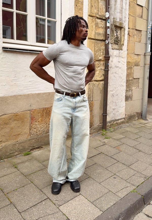 Homme avec dreadlocks portant un t-shirt gris, un jean clair ample et des chaussures noires, debout dans une rue pavée à côté de murs en pierre et en plâtre.