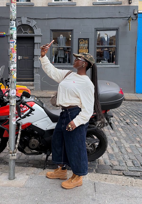 Pitkät letit omaava nainen, joka käyttää maastokuvioista lippalakkia ja saappaita, ottaa selfien punaisen ja valkoisen moottoripyörän luona mukulakivikadulla.
