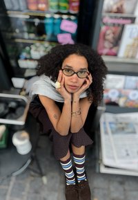 Ung kvinde med krøllet hår og briller, der sidder på en stol med hænderne på ansigtet, iført stribede sokker og et sølvfarvet sjal, omgivet af magasiner.