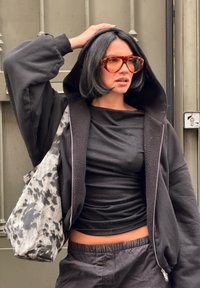 Sort hættetrøje med lynlås og stor hætte, lagdelt over en sort tætsiddende top og sorte bukser. Grå taske med sort abstrakt mønster. Ravfarvede solbriller.