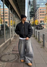 Jeune personne portant un bonnet noir, des lunettes, une veste bomber noire, un jean large délavé et des bottes couleur tan, marchant sur un trottoir urbain près de bâtiments modernes.