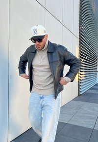 Μαύρο δερμάτινο μπουφάν, ανοιχτό μπεζ πουλόβερ με ψηλό λαιμό, ανοιχτό μπλε φαρδιά τζιν, λευκό καπέλο με λογότυπο και μαύρα γυαλιά ηλίου. Ρυθμίζει το μπουφάν.