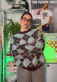 Oversized genser i lys teal og brunt med et argyle-mønster, med rund hals og ribbestrikkede mansjetter, kombinert med nøytrale bukser.