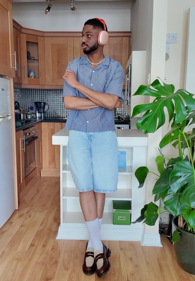 Hombre que lleva una camiseta de rayas azul de manga corta y pantalones cortos de mezclilla deslavados, combinados con calcetines blancos y mocasines marrones; auriculares de cancelación de ruido en color rosa.