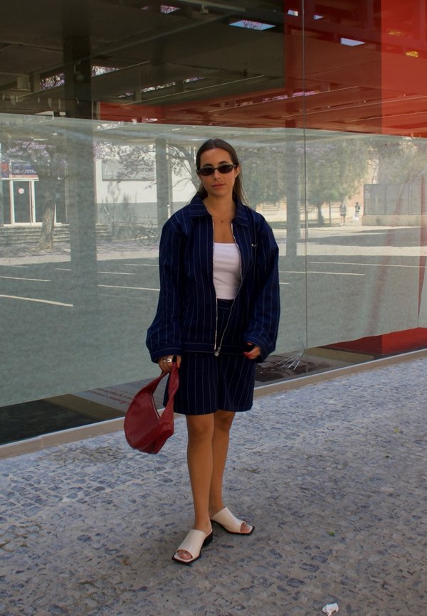 Una mujer lleva una chaqueta oversized de raya diplomática en azul marino y pantalones cortos a juego, combinada con una blusa blanca, sandalias blancas y un bolso rojo.