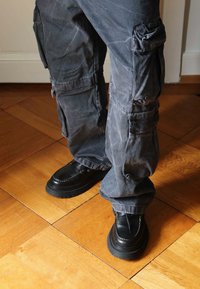 Pantaloni cargo grigi con multiple tasche e vestibilità comoda, abbinati a scarpe in pelle nere con suole spesse e dettagli di cuciture bianche.