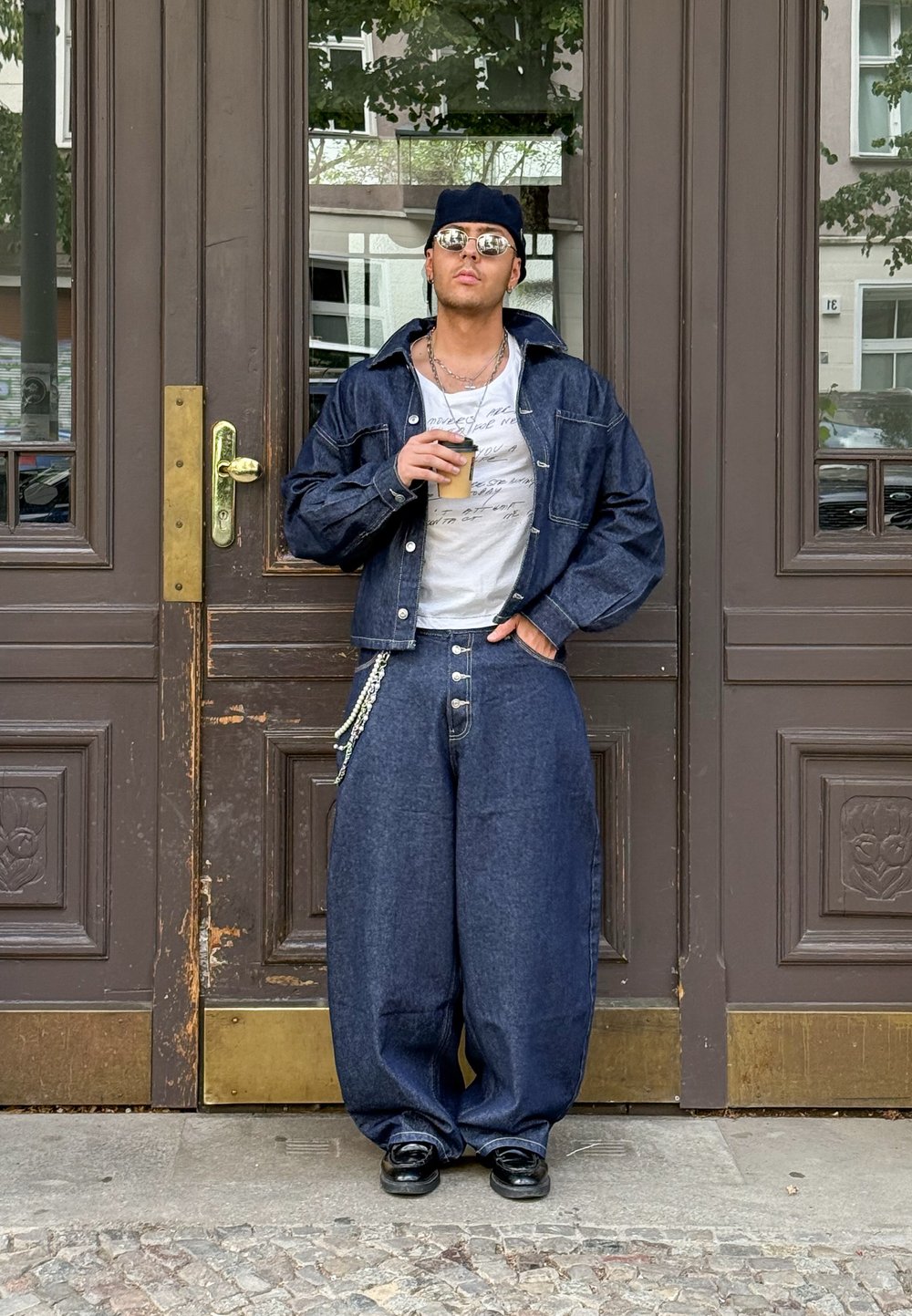 Denim obleka, ki vključuje ohlapen jopič in široke hlače temnega pranja. Bela majica s tekstom, črne čevlje in sončna očala. Drži pijačo.