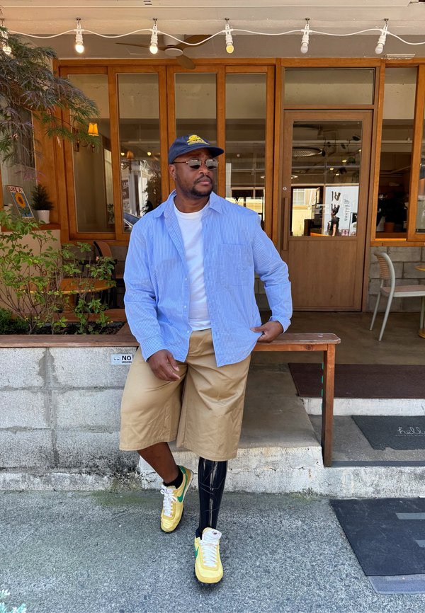 Homme portant une chemise bleue, un short beige, des baskets jaunes et une jambe prothétique noire assis sur un banc en bois devant un café.