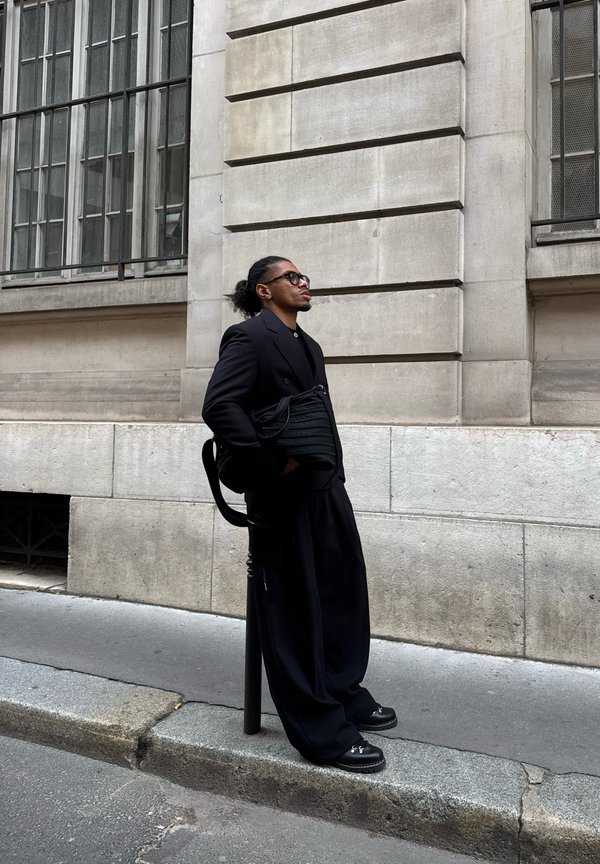 Junger Mann in komplett schwarzem Outfit und Brille, der an einem Pfosten auf dem Bürgersteig einer Stadt an einem steinernen Gebäude mit großen Fenstern lehnt.