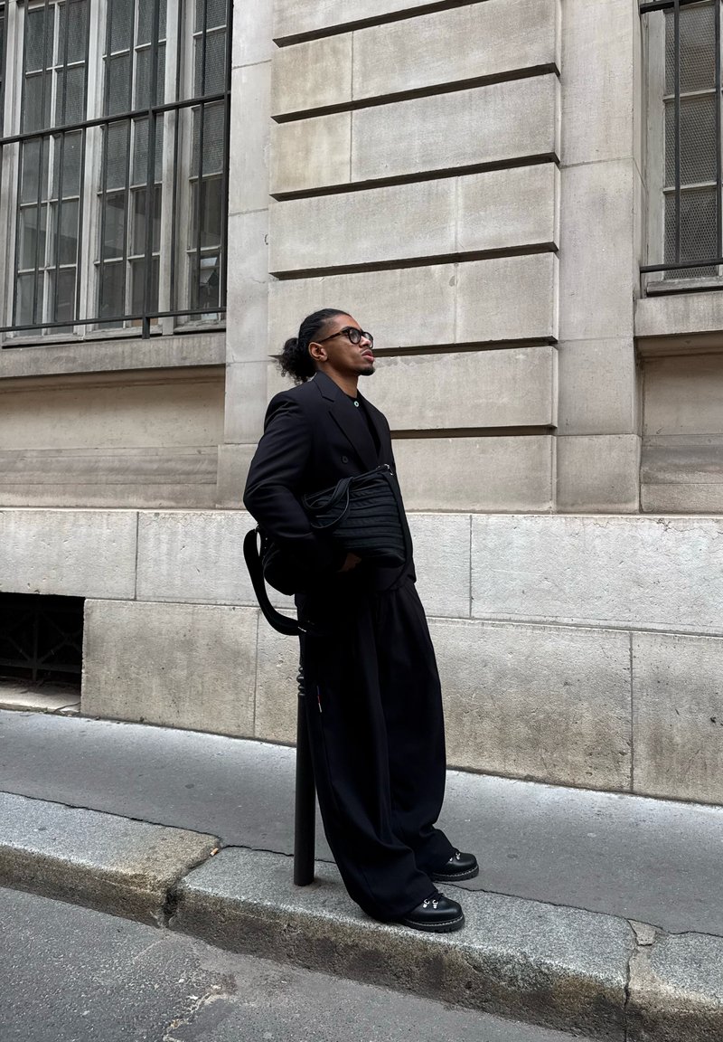 Jeune homme en tenue entièrement noire et lunettes, appuyé contre un poteau sur un trottoir de la ville à côté d'un bâtiment en pierre avec de grandes fenêtres.