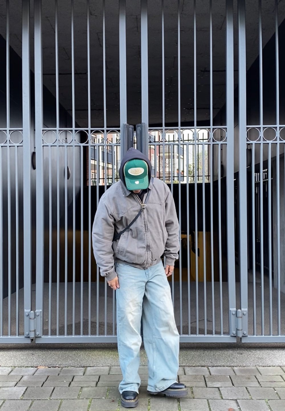 Una persona con una giacca grigia, jeans blu e un berretto verde si trova di fronte a barre metalliche verticali. Il pavimento è lastricato con grandi pietre rettangolari.