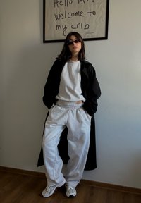 Un sweat-shirt gris et un pantalon de jogging assorti combinés à un long manteau noir. Les chaussures sont des baskets blanches. Du texte encadré est visible sur le mur.