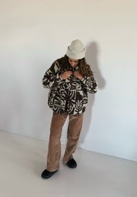 Veste polaire avec un motif tribal audacieux blanc et marron, associée à un pantalon en velours côtelé marron et des chaussures noires sans lacets. Bonnet beige sur la tête.