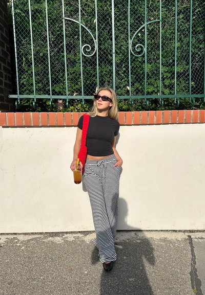 Camiseta negra ajustada, pantalones de pierna ancha a rayas en blanco y negro, bolso de lana rojo, gafas de sol y una taza con una bebida. Fondo de follaje verde.