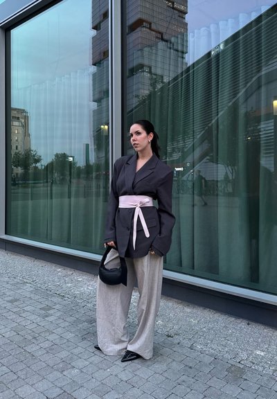 Blazer gris oversized con mangas largas y cinturón rosa, combinado con pantalones de lino beige holgados y un bolso negro, contra un fondo de cristal.