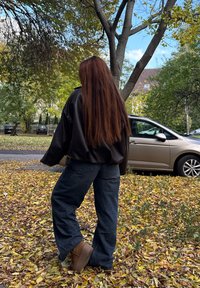 Una persona con lunghi capelli rossi, vestita con una giacca nera e jeans, si trova su foglie autunnali vicino a un albero e a un'auto parcheggiata.