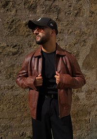 Chaqueta de cuero marrón con corte recto, puños abotonados y cierre de cremallera. Combinada con una camiseta negra y pantalones oscuros frente a una pared texturizada.