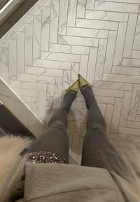 Pelēkas adītas legginses apvienotas ar norādītām, gaiši zaļām kurpēm ar jostas detaļu. Fons rāda marmora rakstu grīdu.