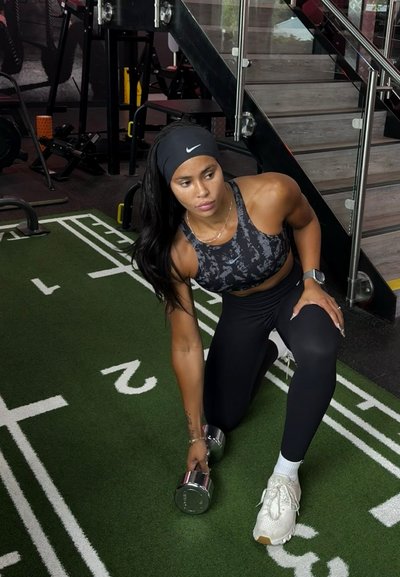 Mancuernas con extremos metálicos circulares, sostenidas en una mano sobre un suelo de gimnasio de césped verde, con ropa de entrenamiento negra y un sujetador deportivo de estampado.
