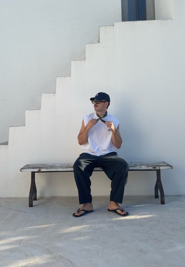 Hombre con gorra, gafas de sol, camiseta sin mangas blanca y pantalones negros sentado en un banco de madera contra una pared blanca con escaleras, ajustando un pañuelo en el cuello.