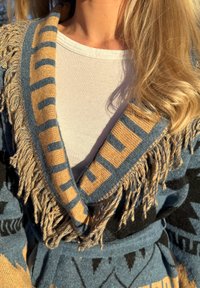 Cardigan tricoté en bleu et beige, avec un motif géométrique et des détails à franges sur le col et les manches. Texture douce, coupe décontractée.