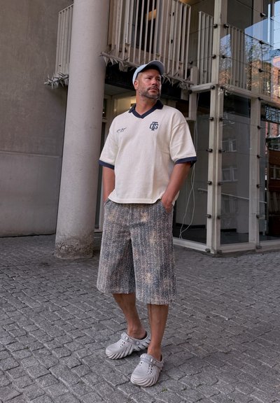 Polo blanco texturizado con acentos en navy, pantalones cortos con estampado y zapatos gris de slip-on. De pie frente a un fondo urbano moderno.