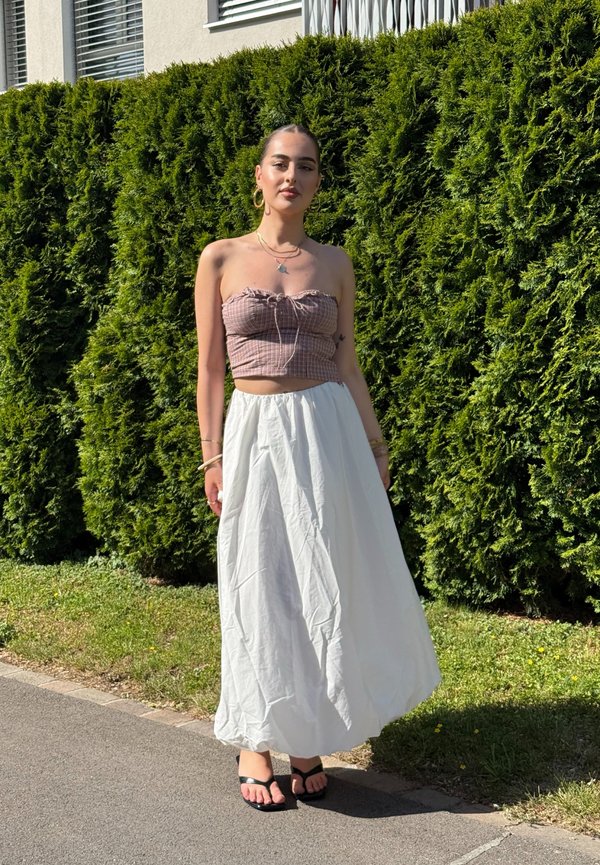 Eine junge Frau steht draußen, trägt ein trägerloses kariertes bauchfreies Top, einen langen weißen Rock, schwarze Sandalen, goldene Creolen und Armbänder vor grünem Heckenhintergrund.