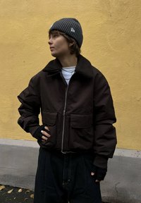 Smeđa bomber jakna s ovratnikom od ovčje vune, dva prednja džepa, nošena preko bijele košulje, uparena sa sivom pletenom kapom i tamnim hlačama.