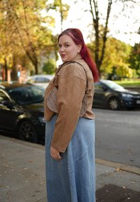 Kurtka zamszowa w kolorze jasnobrązowym, niebieska długa spódnica dżinsowa z widocznymi przeszyciami oraz beżowa torba na ramię z wzorem. Jasnoczerwone włosy, na zewnątrz w otoczeniu drzew.