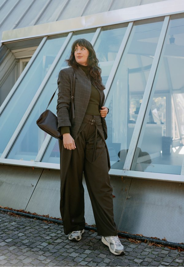 Femme aux longs cheveux foncés portant un blazer marron à carreaux, un pantalon foncé et des baskets, se tient devant un immeuble moderne en verre avec un sac à bandoulière marron.