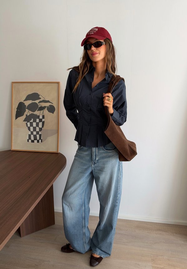 Mujer con gorra roja, gafas de sol negras, camisa de cuadros oscura, vaqueros anchos azul claro, bolso marrón de hombro, de pie junto a una mesa de madera con arte de plantas enmarcado.