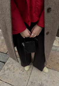 Personne portant un manteau marron, une veste rouge, un pantalon foncé et des chaussures claires, tenant un petit sac à main noir à deux mains.