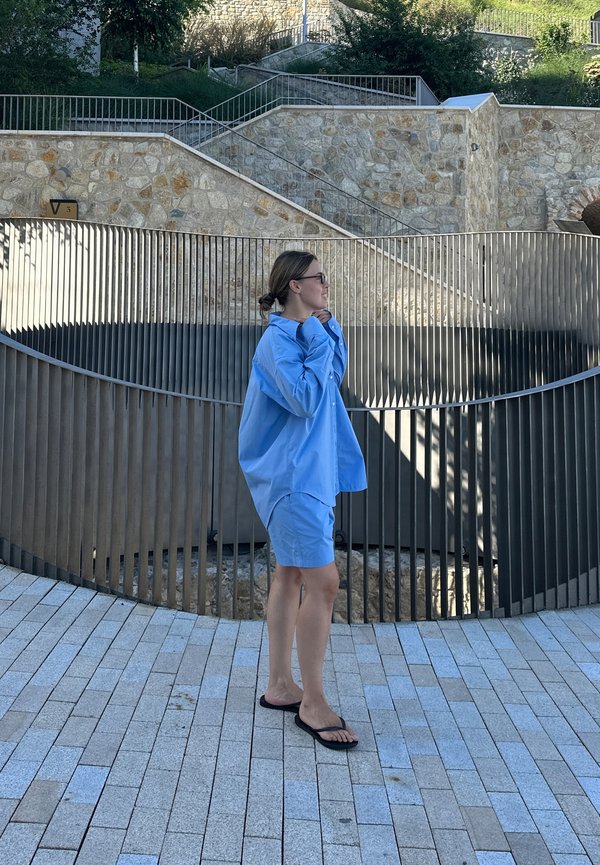 Camisa azul clara oversized com botões, combinada com calções iguais. O tecido parece suave, complementado por chinelos pretos.