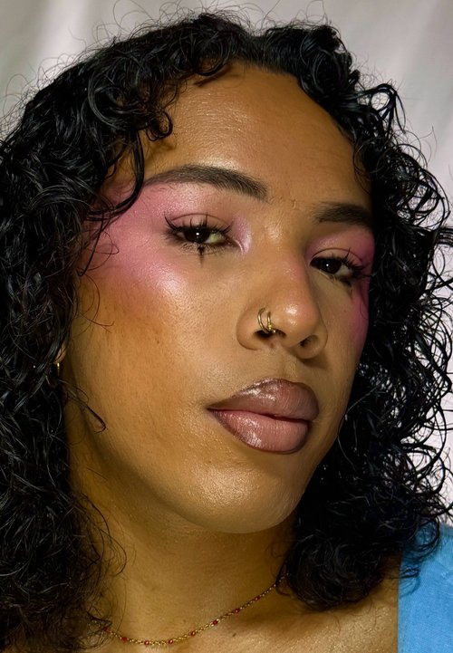 Person mit lockigem schwarzen Haar, rosa Lidschatten, glänzenden Lippen, goldenen Nasenringen und einer zarten Perlenkette vor hellem Hintergrund.