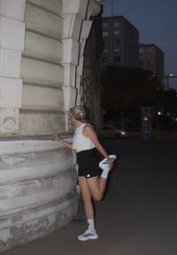 Persona atlética estirándose cerca de una columna de piedra texturizada. Lleva un crop top blanco, pantalones cortos negros, zapatillas deportivas blancas y azules, y una gorra con un patrón.