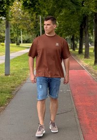 Brązowa koszulka z krótkim rękawem, niebieskie szorty dżinsowe i szare sneakersy; modelka idąca ścieżką otoczoną zielenią.