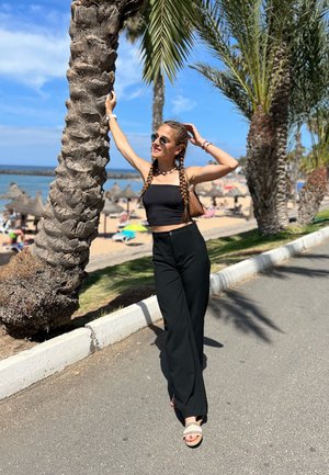 Top corto negro y pantalones anchos negros. La persona lleva gafas de sol y está parada al lado de una palmera, con una playa de fondo.