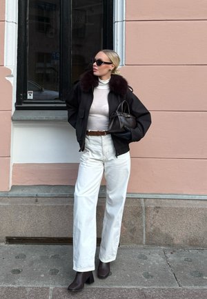 Mujer con chaqueta negra con cuello de piel, suéter de cuello alto blanco, pantalones blancos, botas negras, gafas de sol, sosteniendo un bolso negro, de pie junto a una pared y ventana rosa.