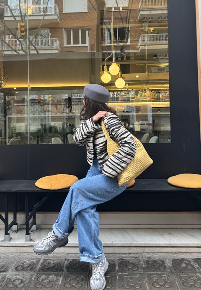 Chaqueta acolchada a rayas, boina gris claro, bolso tejido amarillo, jeans azules holgados y zapatillas grises. Sentado en un banco negro con cojines redondos.