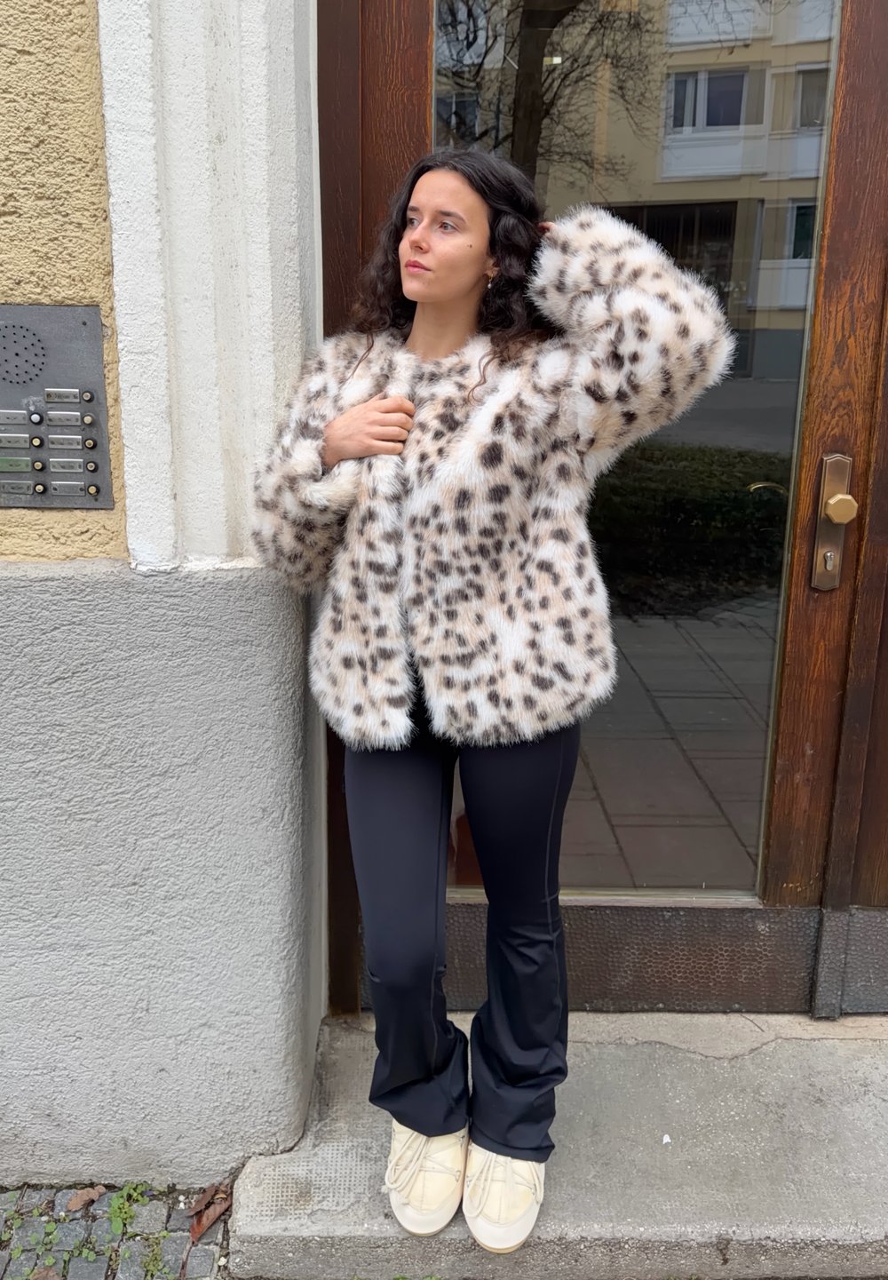 Jakna iz umetnega krzna z leopardjim vzorcem, v krem in rjavih tonih, sproščenega kroja; kombinirana s črnimi oprijetimi hlačami in kremnimi čevlji.